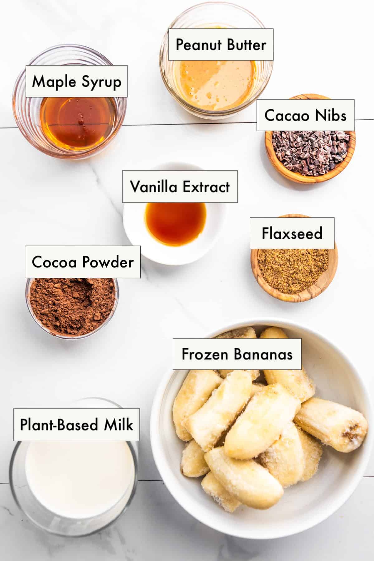 All ingredients for a banana cocoa peanut butter smoothie in labeled bowls on a counter.
