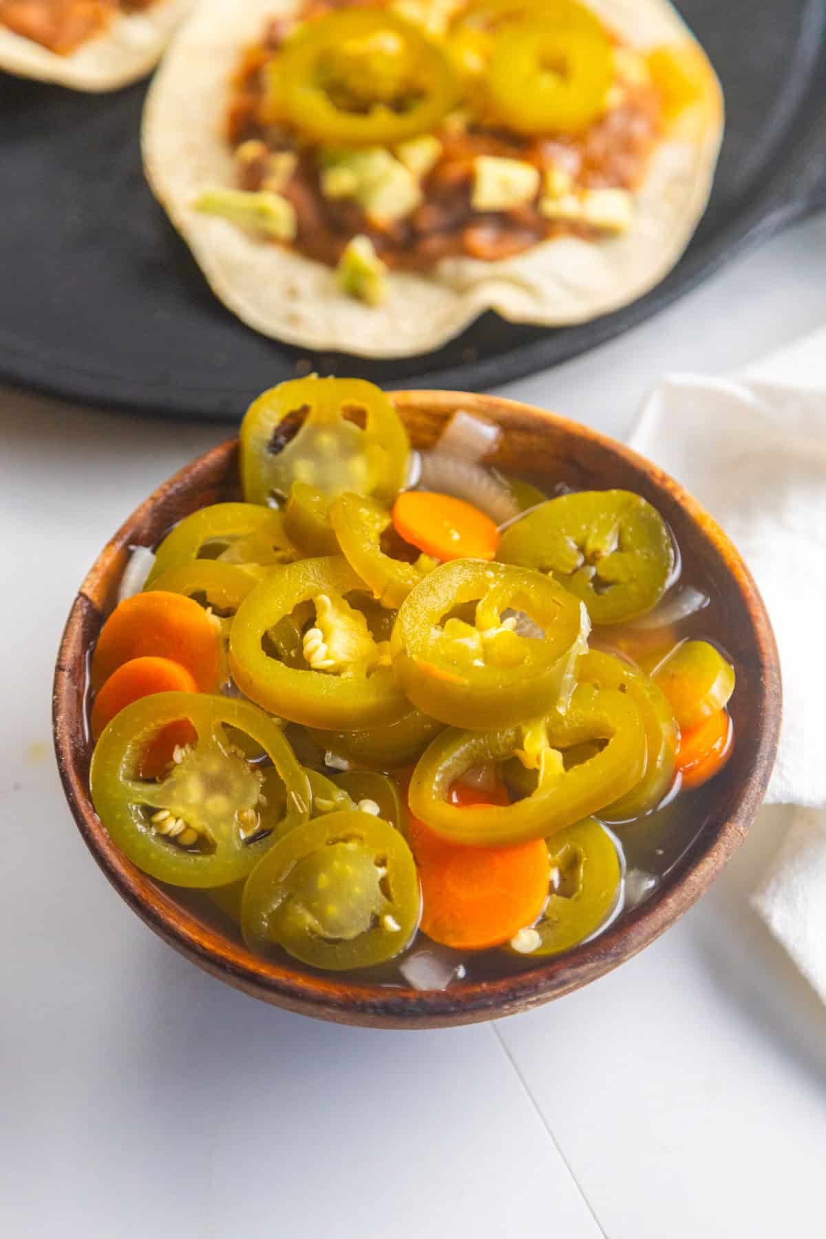 Pickled jalapeños in a wooden bowl with homemade tacos in the background.
