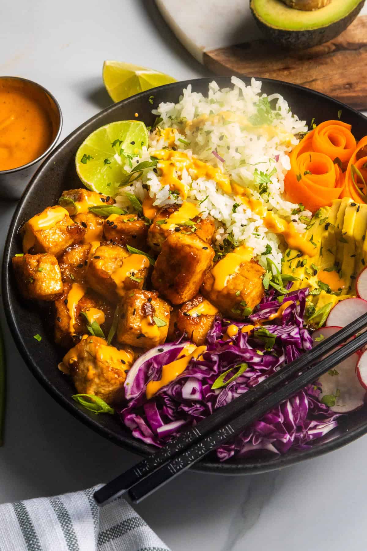 Sticky tofu bowl (AKA crispy tofu rice bowl) in a large black bowl with a dipping sauce and sliced avocado on the side.