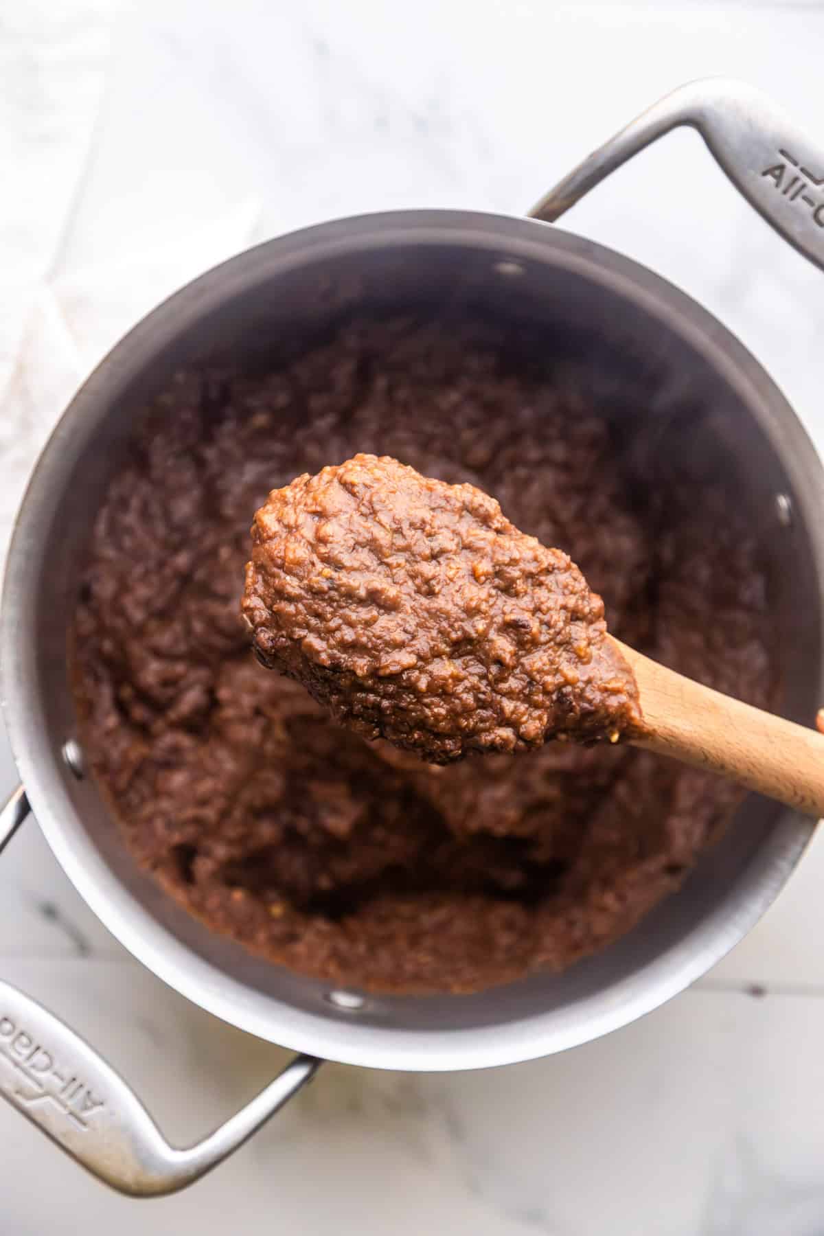Vegan refried black beans from canned beans in a pot being scooped up with a spoon.