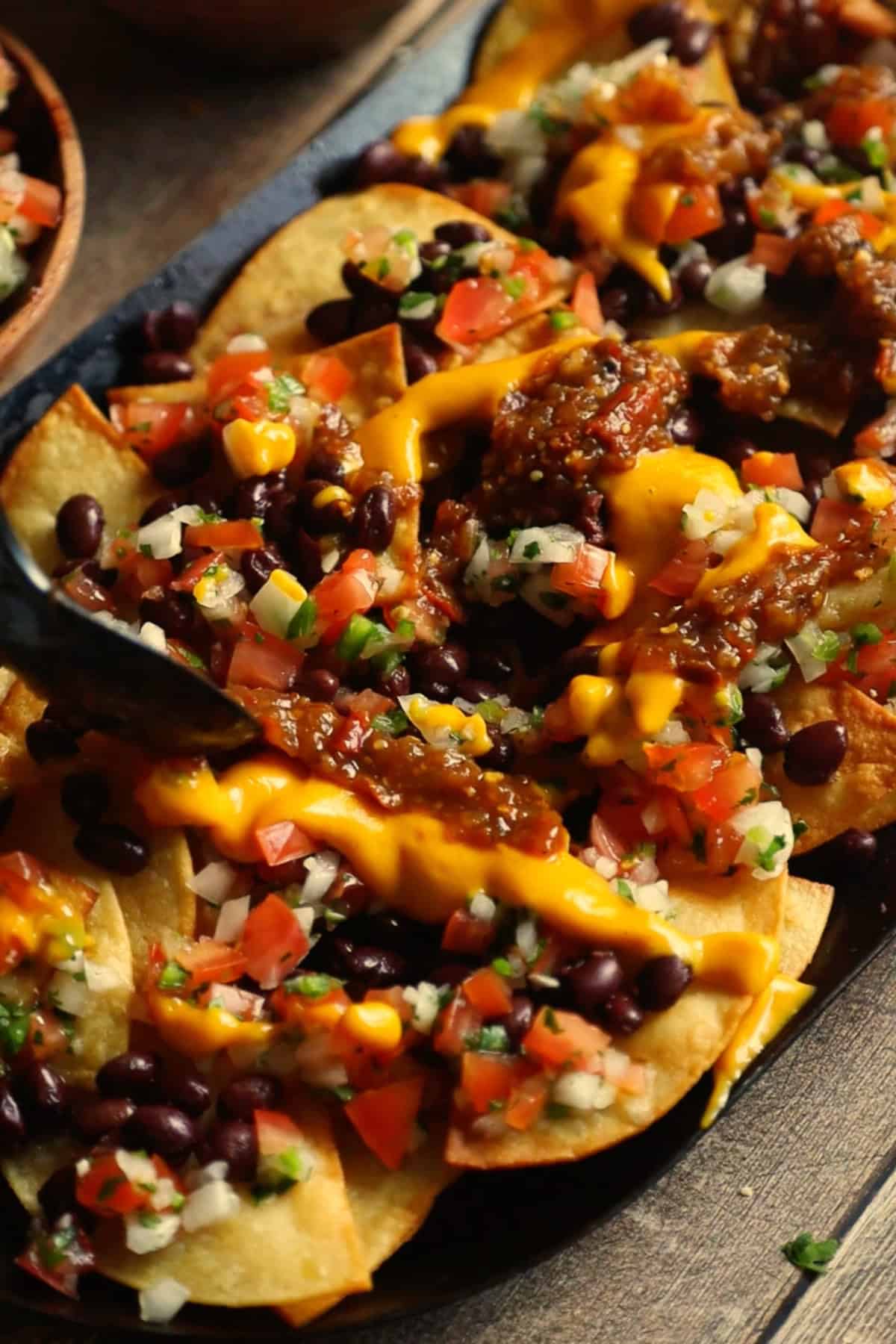 Vegan loaded nachos on a comal being topped with pico de gallo, cheese sauce, salsa, and guacamole.