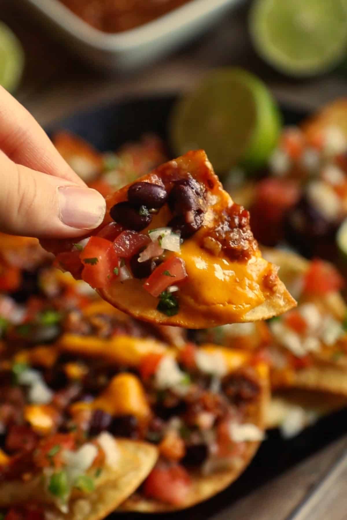 A hand taking a loaded nacho chip from a plate of vegan nachos covered in guacamole, pico de gallo, and salsa.